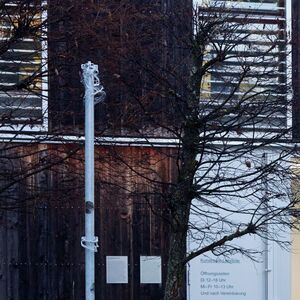 Eine schlanke Metallstele steht auf nassem Gehweg vor einem modernen Gebäude mit Holz- und Glasfassade, grünem Rasen und einem kahlen Baum.