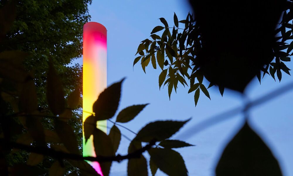 Leuchtende, vertikale Lichtskulptur in Regenbogenfarben, umgeben von Bäumen und Sträuchern.