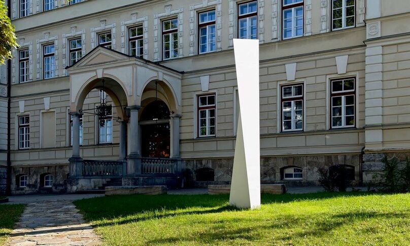 Eine schlanke, weiße Skulptur steht auf einer grünen Rasenfläche vor einem historischen Gebäude mit hohen fenstern und Rundbogeneingang.