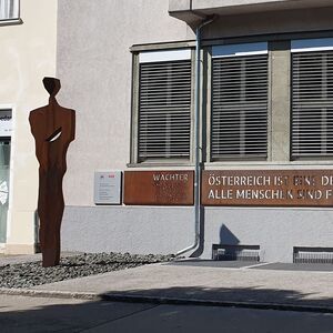 Links steht eine bronzefarbene Metallskulptur vor einem Gebäude, an der Fassade ist ein bronzener Schriftzug befestigt