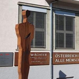 Bronzene Metallfigut steht vor einem grauen Gebäude, an der Fassade ist ein bronzefarbener Schriftzug befestigt.