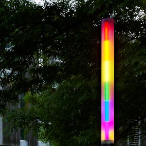 Leuchtende, vertikale Lichtskulptur in Regenbogenfarben im Außenraum vor einem modernen Gebäude, umgeben von Bäumen und Sträuchern.