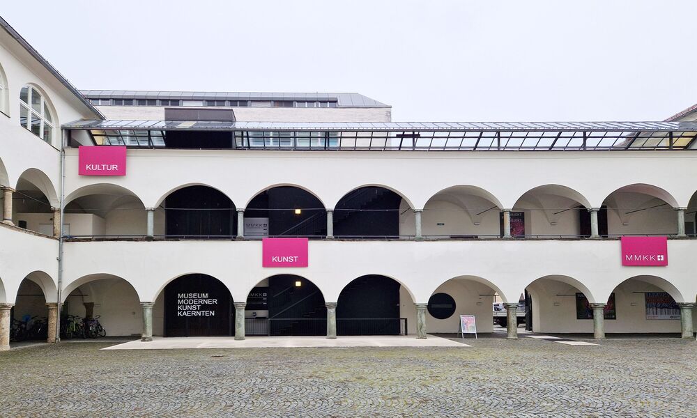 Heller, zweigeschossiger Arkadenhof mit Rundbögen, gepflastertem Boden und mehrere pinkfarbenen Schildern, die auf das Museum Moderner Kunst Kärnten, sowie auf die Kulturabteilung hinweisen.