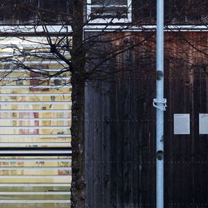 Eine schmale Metallstele steht auf einer nassen Gehfläche vor einem modernen Gebäude mit Holz- und Glasfassade, einem Baum und grünem Rasen.