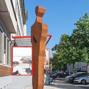 Bronzefarbene Metallskulptur steht zwischen einem Gebäude und einem Gehweg