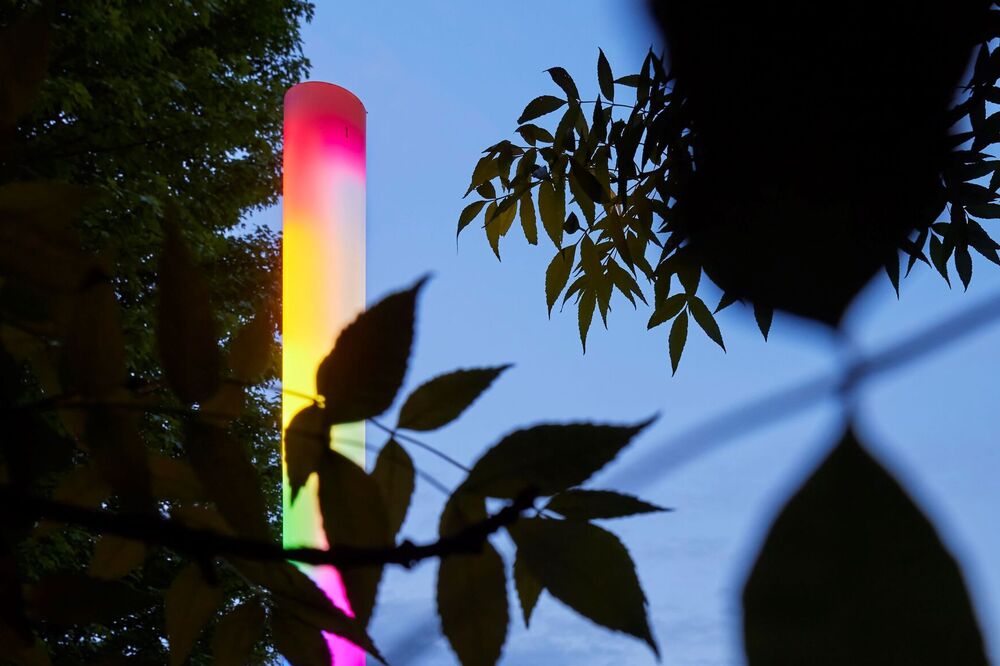 Leuchtende, vertikale Lichtskulptur in Regenbogenfarben, umgeben von Bäumen und Sträuchern.