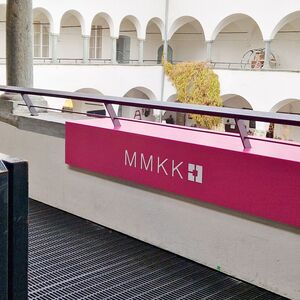 Blick in einen hellen Arkadenhof mit schwarzer Treppe und einem pinkfarbenen MMKK-Schild im Geländer