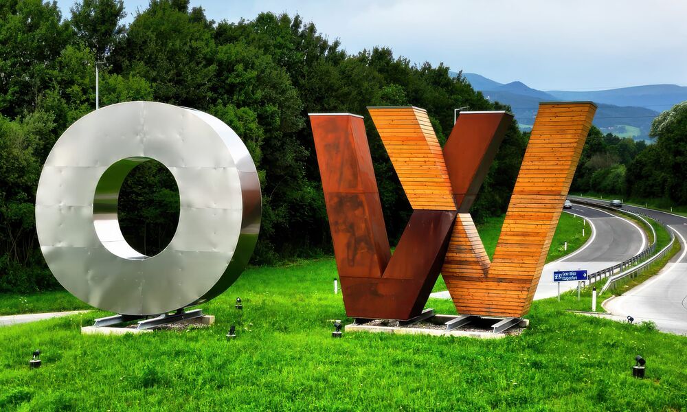 Die Buchstaben O und W als große Skulpturen aus Metall und Holz auf einer grünen Wiese neben einer Autobahnauffahrt