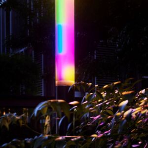 Leuchtende, vertikale Lichtskulptur in Regenbogenfarben, umgeben von Bäumen und Sträuchern.