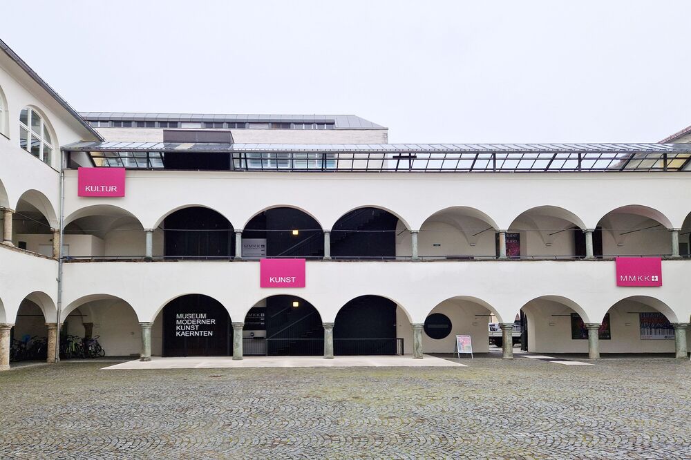 Heller, zweigeschossiger Arkadenhof mit Rundbögen, gepflastertem Boden und mehrere pinkfarbenen Schildern, die auf das Museum Moderner Kunst Kärnten, sowie auf die Kulturabteilung hinweisen.