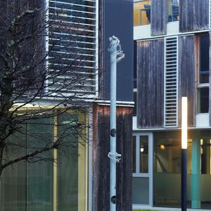 Das Bild zeigt einen modernen Gebäudekomplex mit Holz- und Glasfassaden, davor eine Stele mit der Aufschrift "B02", nassem Gehweg, etwas Rasen und einem kahlen Baum bei feuchtem Wetter.