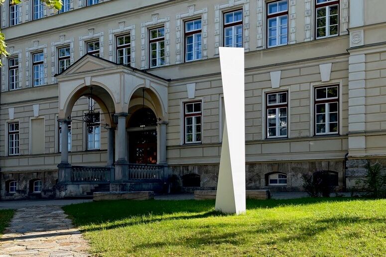 Eine schlanke, weiße Skulptur steht auf einer grünen Rasenfläche vor einem historischen Gebäude mit hohen fenstern und Rundbogeneingang.